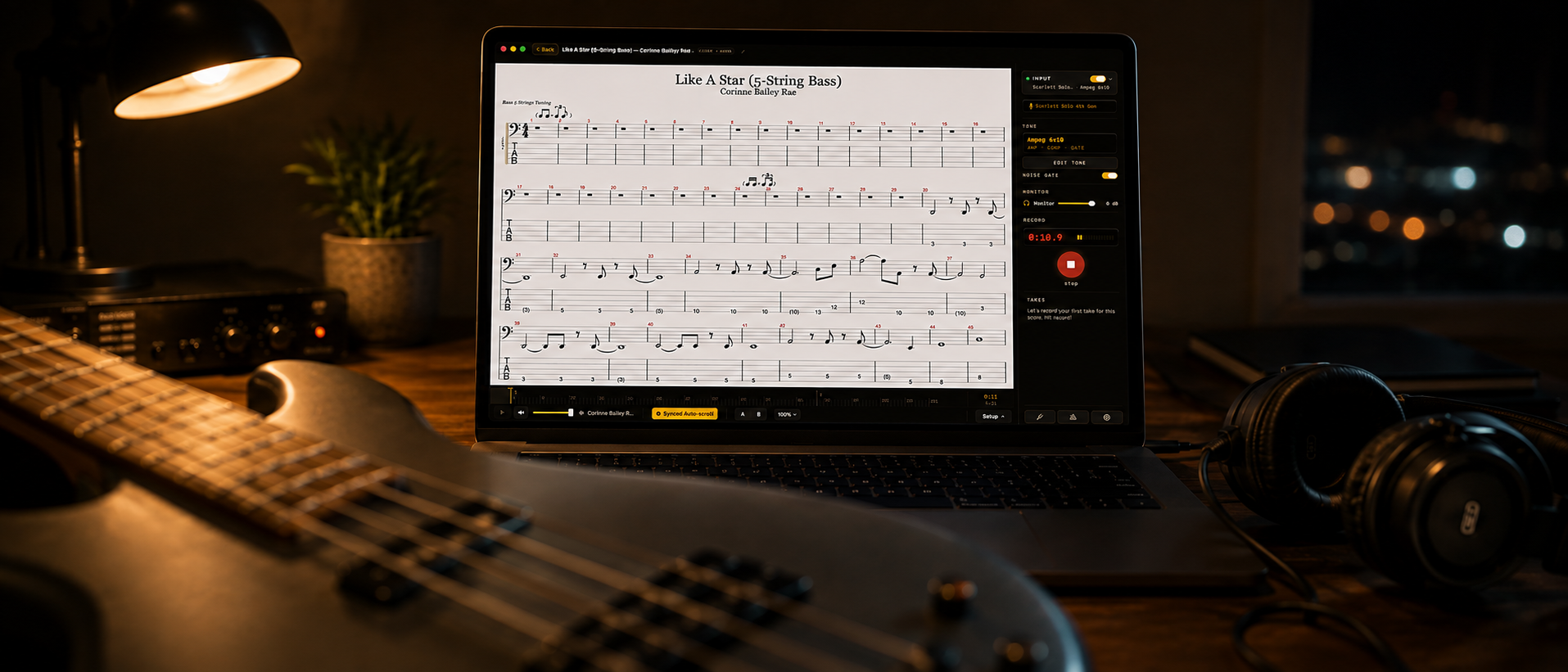 A MacBook Pro at night on a wooden desk, showing a five-string bass score for Like A Star by Corinne Bailey Rae in Afinar, with Synced Auto-scroll lit and the recorder mid-take. A bass lies across the desk in the foreground, with a lamp, an interface and headphones around it.
